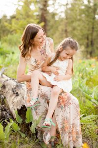 mother in floral dress tickling daughter in white as they sit on a log