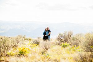 wide image of man holding pregnant wife from behind on flower covered hillside